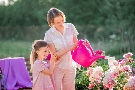 Mom and daughter in a linen pink dress take care of peonies in the garden at the cottage, water the flowers. He perfectly smiles and laughs. Illuminated by the rays of the evening setting sun.の写真素材