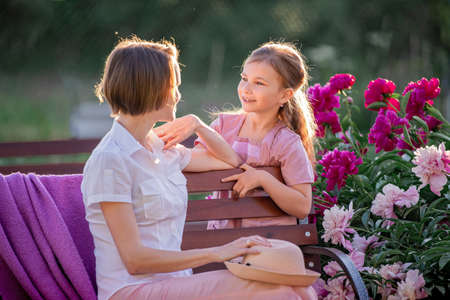 Happy family relationships. Mom and daughter sit on a bench and chat in the garden among the flowers on a sunny summer evening.の写真素材