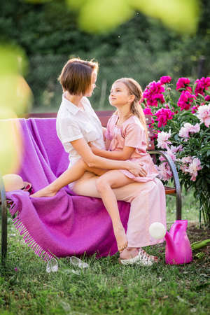 Happy family relationships. Mom and daughter sit on a bench and chat in the garden among the flowers on a sunny summer eveningの写真素材