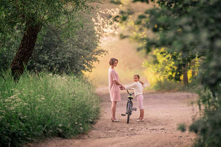 mom and daughter are walking summer evening in the sun in a park with a bicycleの写真素材