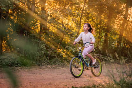 little girl goes with a bicycle on a rural road in nature at sunsetの写真素材