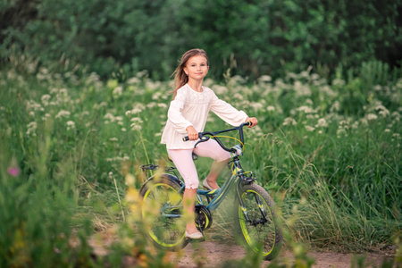 ittle girl rides a bicycle on a rural road in nature in a park among tall wildflowers. Rushing fast, hair fluttering in the wind.の写真素材
