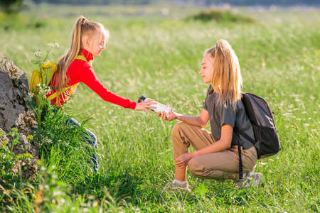 Children with backpacks on a hike on a summer trip to nature. One girl passes another bottle of water.の写真素材
