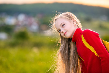 portrait of a child girl in a red jumper with a backpack traveling on a summer evening against the backdrop of a landscape.の写真素材