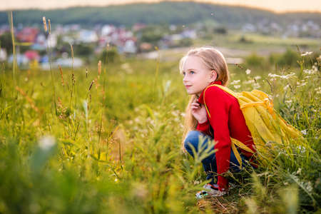 portrait of a child girl in a red jumper with a backpack traveling on a summer evening against the backdrop of a landscape.の写真素材