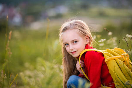 portrait of a child girl in a red jumper with a backpack traveling on a summer evening against the backdrop of a landscape.の写真素材