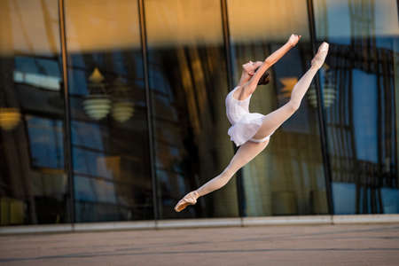 a young ballerina in a white leotard is dancing on pointe shoes against the background of a cityscape, frozen in a jumpの写真素材