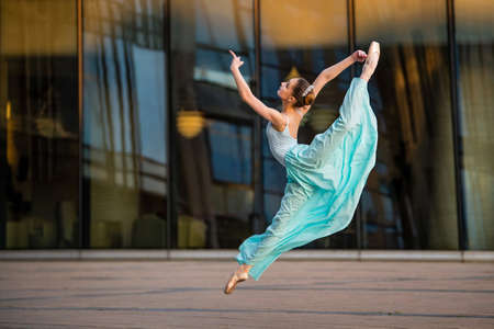 a young ballerina in a white leotard is dancing on pointe shoes against the background of a cityscape, frozen in a jumpの写真素材