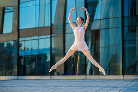 a young ballerina in a white leotard is dancing on pointe shoes against the background of a cityscape, frozen in a jumpの写真素材