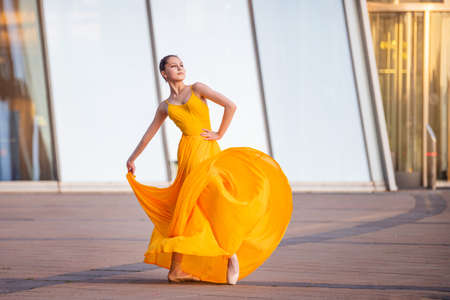 a young ballerina in a long flying yellow dress is dancing against the backdrop of a cityscapeの写真素材