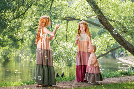 Three red-haired sisters in long linen dresses blow bubbles in the park on a sunny summer day.の写真素材