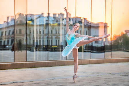 young ballerina is dancing on pointe shoes in a delicate mint blue tutu against the backdrop of the reflection of a sunset in the cityの写真素材