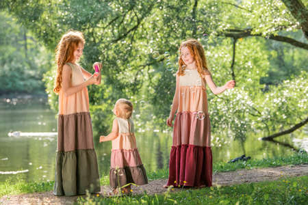 Three red-haired sisters in long linen dresses blow bubbles in the park on a sunny summer day.の写真素材