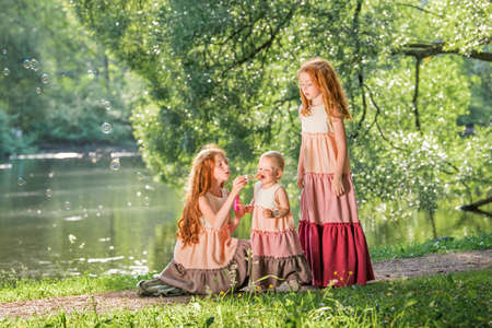 Three red-haired sisters in long linen dresses blow bubbles in the park on a sunny summer day.の写真素材