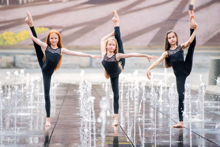 a group of three little ballerinas in black tight-fitting suits dance against the backdrop of city fountains on a hot day.の写真素材