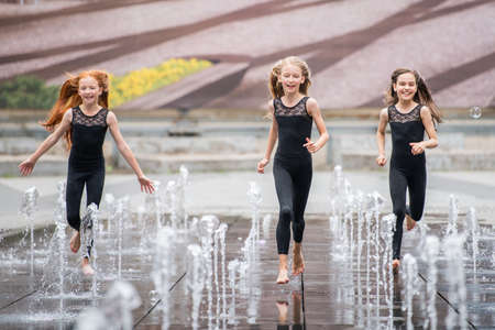group of three little ballerinas in black tight-fitting suits run to the viewer amid splashing fountainsの写真素材