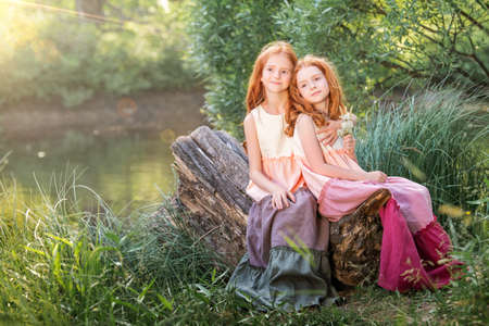 Two red-haired sisters in long linen dresses are resting on the lake in the park on a sunny summer day.の写真素材