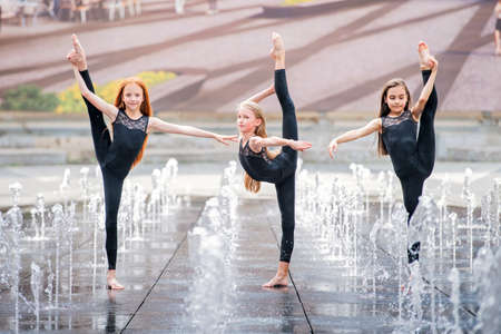 a group of three little ballerinas in black tight-fitting suits dance against the backdrop of city fountains on a hot day.の写真素材
