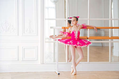Portrait of a little full-length ballerina in a bright crimson tutu standing at the machine in front of a mirrorの写真素材