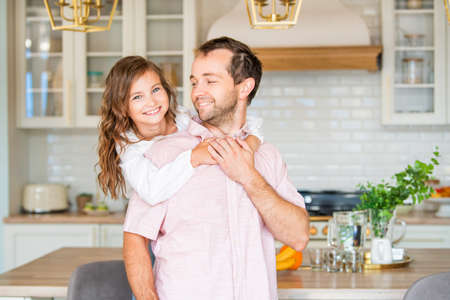 Cute little girl in casual clothes hugs her handsome father at home in the kitchen. Both look at the camera and smile.の写真素材