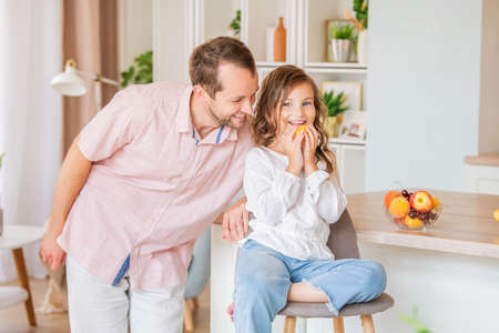 Cute little girl in casual clothes eats a peach at home in the kitchen, her happy father hugs her.の写真素材