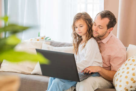 Happy family, daughter sitting on her father's lap on the couch at home looking at the laptop monitor together.の写真素材