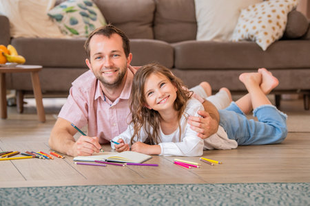 Father and daughter are drawing together while lying on the floor in the apartment room.の写真素材