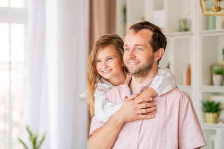 Cute little girl in casual clothes hugs her handsome father at home in the kitchen. Both look at the camera and smile.