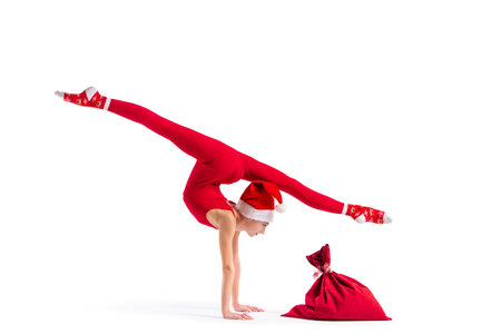 Slender girl gymnast in red tight-fitting suit and santa claus hat posing next to a bag for gifts isolated on white backgroundの写真素材