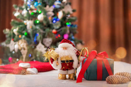 Christmas, Irushechny Santa with a gift box on the background of a Christmas tree and blurry lights of garlands.の写真素材