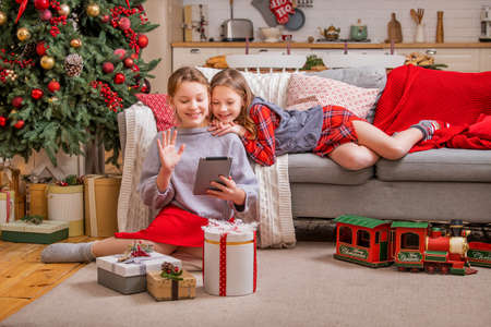 Two joyful sisters are sitting at home near the christmas tree and looking at the tablet monitorの写真素材