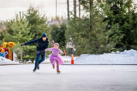 little girl in a pink sweater and a fluffy skirt on sunny winter day rides with her dad on an open skating rink in parkの写真素材