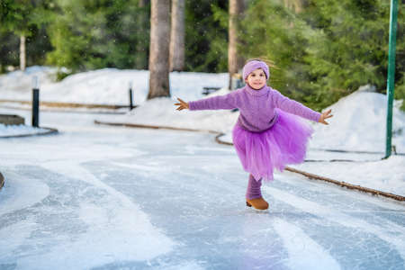 little girl in a pink sweater and a full skirt rides on a sunny winter day on an outdoor ice rink in the parkの写真素材