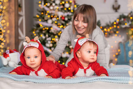 toddler twins in red reindeer santa claus costumes are sitting at home with their mother against the background of the christmas treeの写真素材