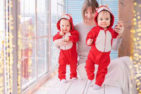 toddler twins in red reindeer santa claus costumes are sitting at home with their mother against the background of a window with garlandsの写真素材