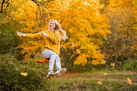Happy young blonde woman with loose hair smiles and rides on rope swing in autumn in park.の写真素材