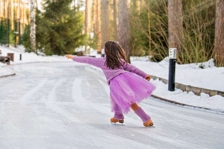 little girl in a pink sweater and a full skirt rides on a sunny winter day on an outdoor ice rink in the parkの写真素材