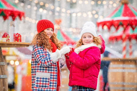 Children, red-haired sisters warm their gloved hands with mug of hot tea at a festively decorated Christmas market in city.の写真素材