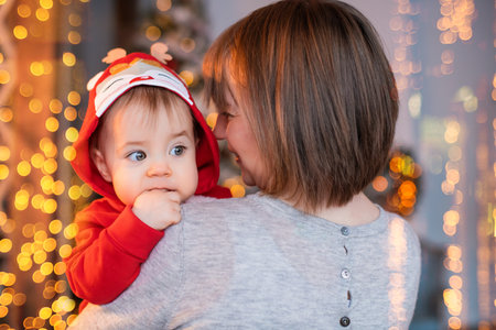 happy mom playing with her toddler in red santa reindeer costume on background of christmas tree and lights garlandsの写真素材