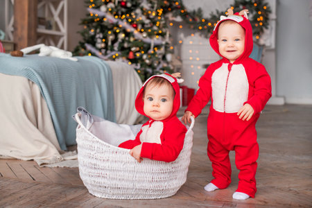 twins toddlers in red santa's reindeer suits sit next to each other at home against the background...