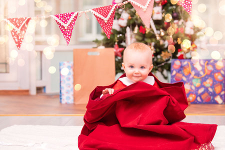 adorable toddler dressed as Santa peeks out of Santa Claus Christmas sack in decorated living room with Christmas tree.の写真素材