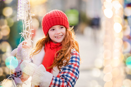 child red-haired girl walk and look at the stall with garlands at festively decorated Christmas market in city.の写真素材