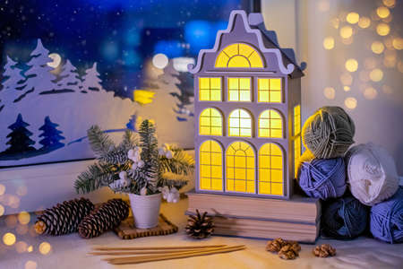On the windowsill, among the skeins of yarn, a night light in the form of an old European house glows against the background of a night snow window and paper decorations.の写真素材