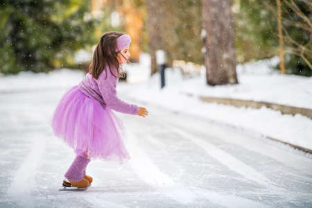 little girl in a pink sweater and a full skirt rides on a sunny winter day on an outdoor ice rink in the parkの写真素材