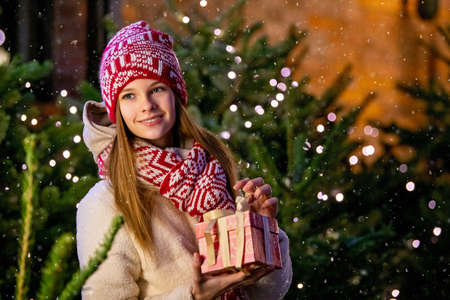 Christmas portrait of a beautiful girl in a warm scarf, standing on the street, decorated for the New Year, against the background of a Christmas tree with a gift in her handsの写真素材