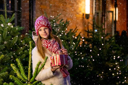 Christmas portrait of a beautiful girl in a warm scarf, standing on the street, decorated for the New Year, against the background of a Christmas tree with a gift in her handsの写真素材