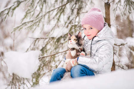 little girl sits with cat in an embrace in the snow in the winter forestの写真素材