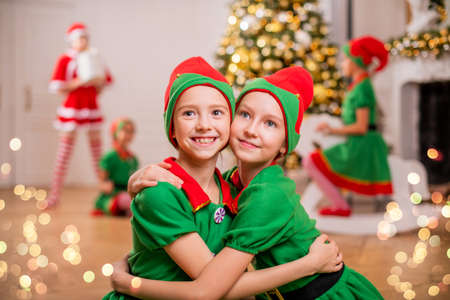 Two cheerful happy girls-friends in costumes of Santa's elves on background of Christmas tree and other childrenの写真素材