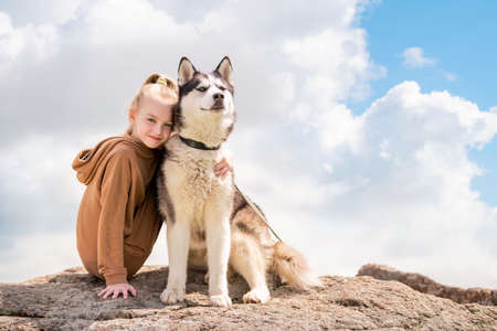Siberian husky and his beautiful blue-eyed mistress are sitting on a rock against sky. The concept of love for animals and nature.の写真素材