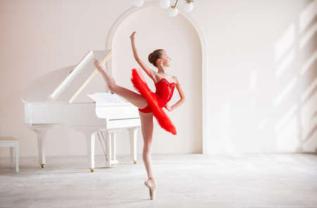 cute little girl dreams of becoming professional ballerina. In a white room, next to a piano, girl in a bright red tutu is dancing on pointe shoes. Vocational school student.の写真素材